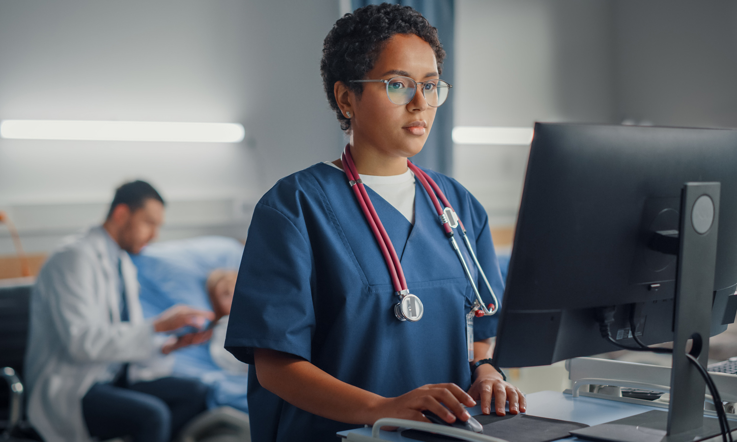 A health care provider uses a computer in a hospital room. A patient and doctor are in the background.